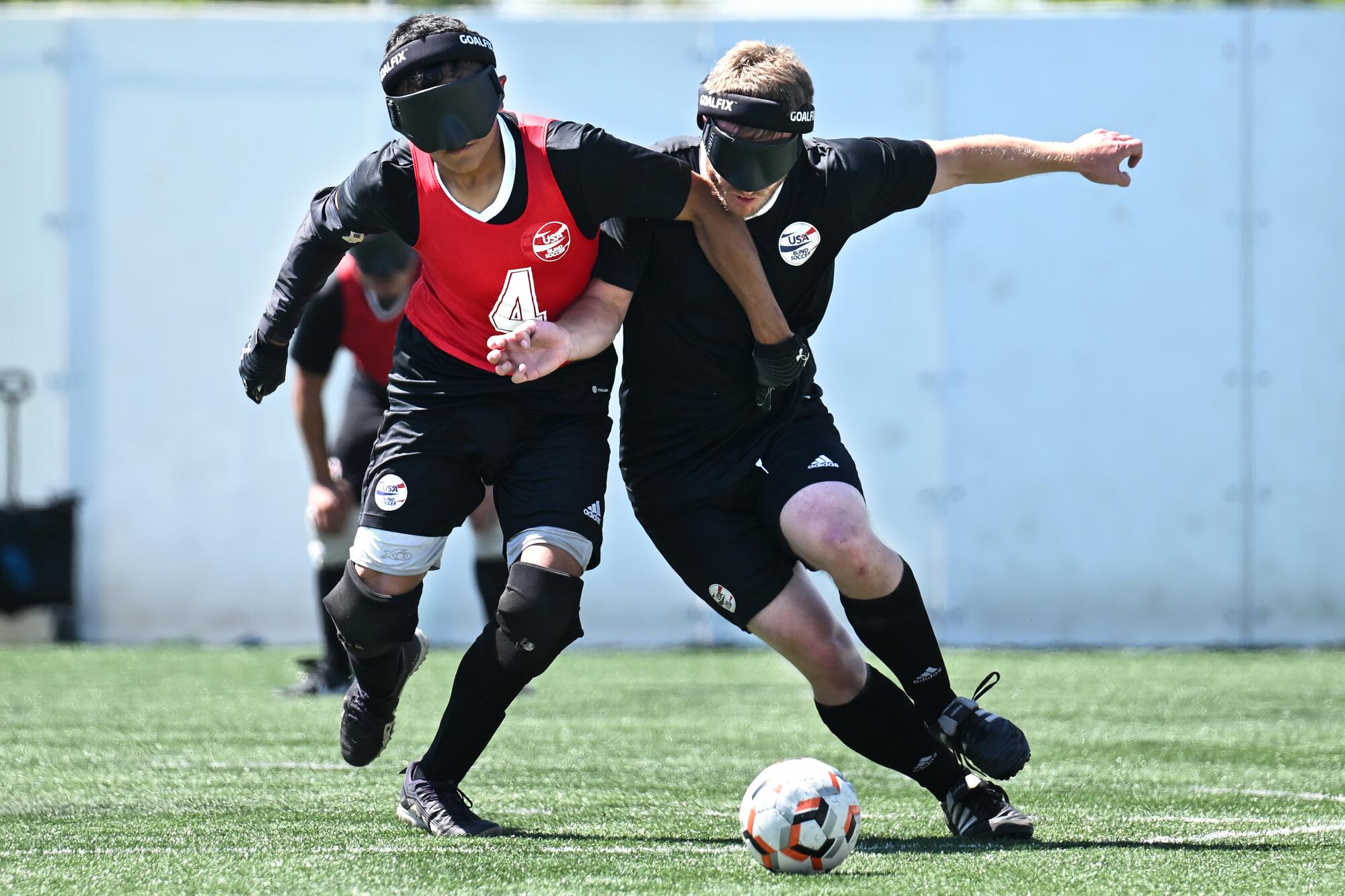 Two blindfolded soccer players in USA Blind Soccer jerseys battling for control of the ball.