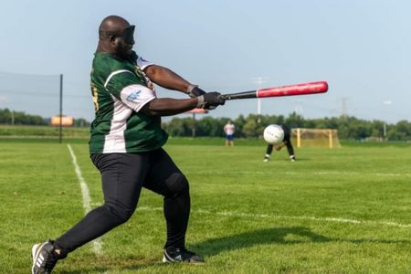 A beep baseball player mid-swing on the field with a catcher ready behind home plate