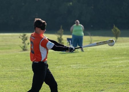 A blindfolded beep baseball batter swings while a spotter watches from the outfield