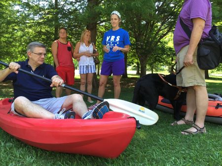 A volunteer explains kayak setup to a group, including a participant with a guide dog