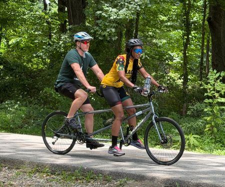 Two tandem cycling riders pedal together along a wooded trail